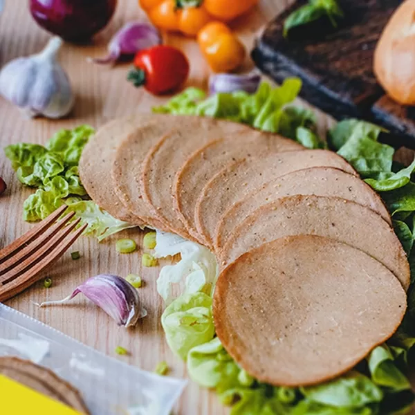 Vegan Garlic Ham Slices by Plenty Reasons shown on a table with garlic, a baguette and salad.