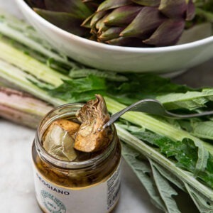 Sicilian Artichoke Hearts Antipasti by Seggiano shown being picked out of the jar surrounded by artichokes and fresh chard.