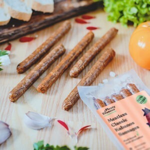 Vegan Classic Kabanos Sausages by Plenty Reasons shown on a table with onions, bread slices and vegetables.