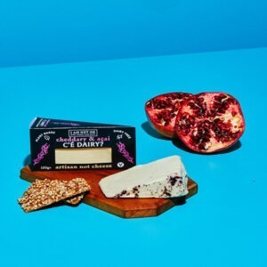 Vegan "C'è Dairy?" Cheddary & Açaí wedge by I Am Nut OK shown on a board with crackers and an open pomegranate.